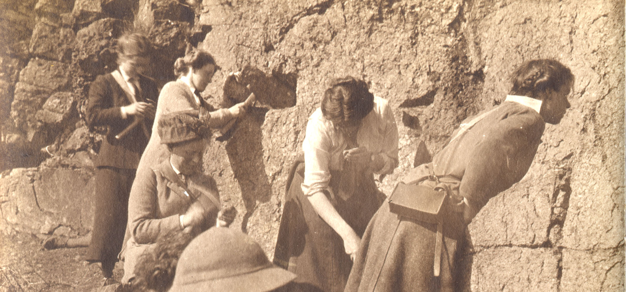  A sepia photograph of 5 female, early geologists studying a rock - face wall, some tapping the wall and rocks with geological hammers.