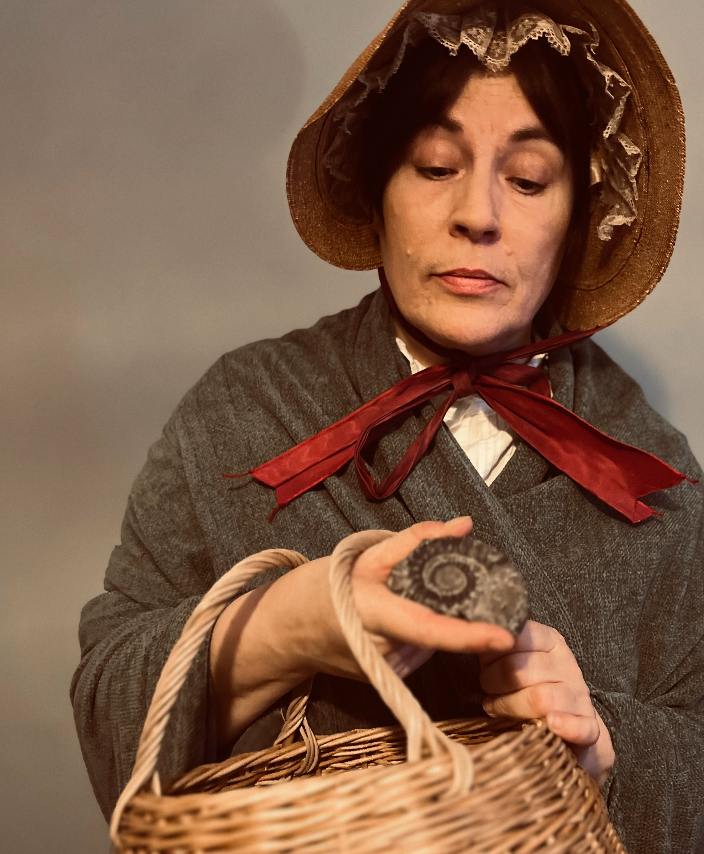 An actress playing the palaeontologist Mary Anning. She is wearing a shawl and bonnet with a red ribbon. She is holding a basket and an ammonite fossil, which she is looking at carefully.