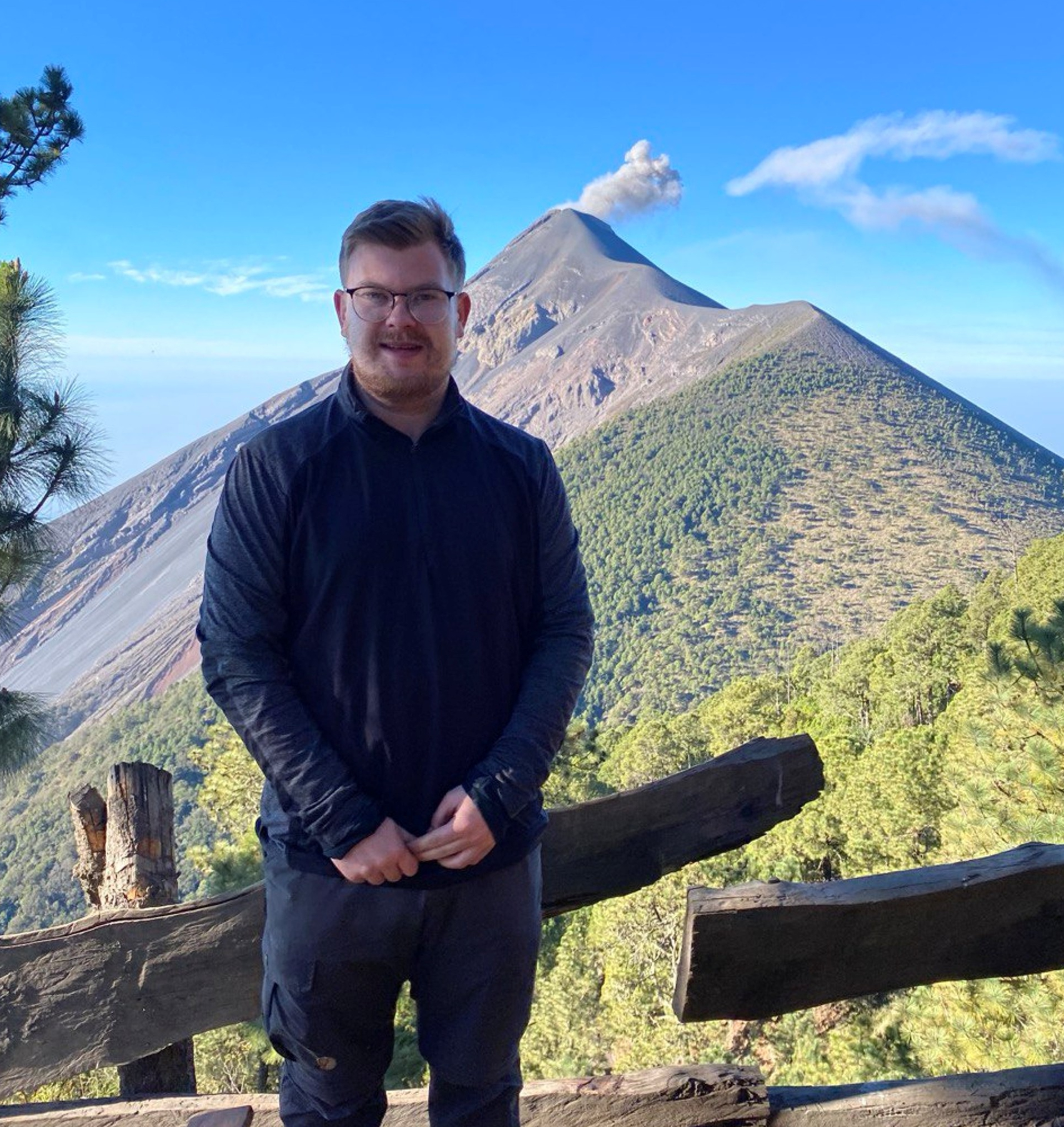 ​​​​Earth Scientist Julian Pahl stands front of a volcano and blue sky. The volcano is smoking slightly.