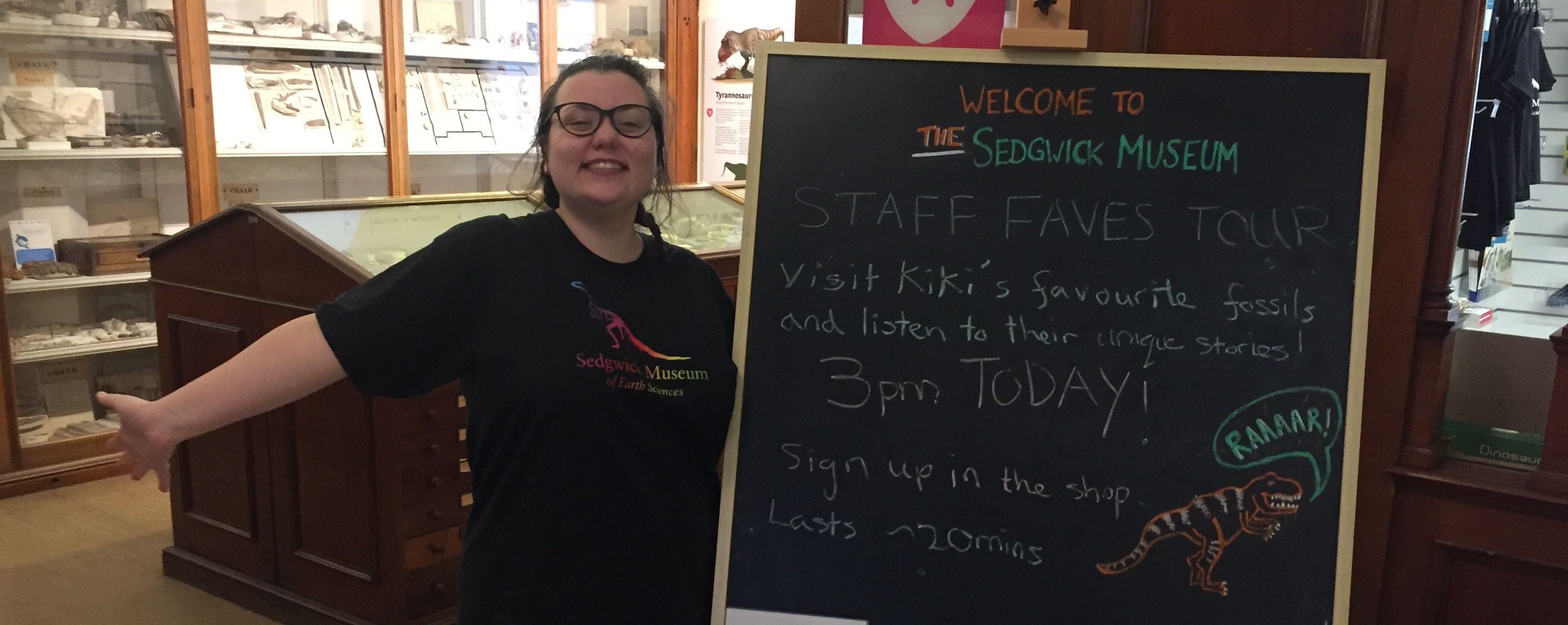 A member of museum staff enthusiastically smiling next to a chalk board sign advertising her tour.