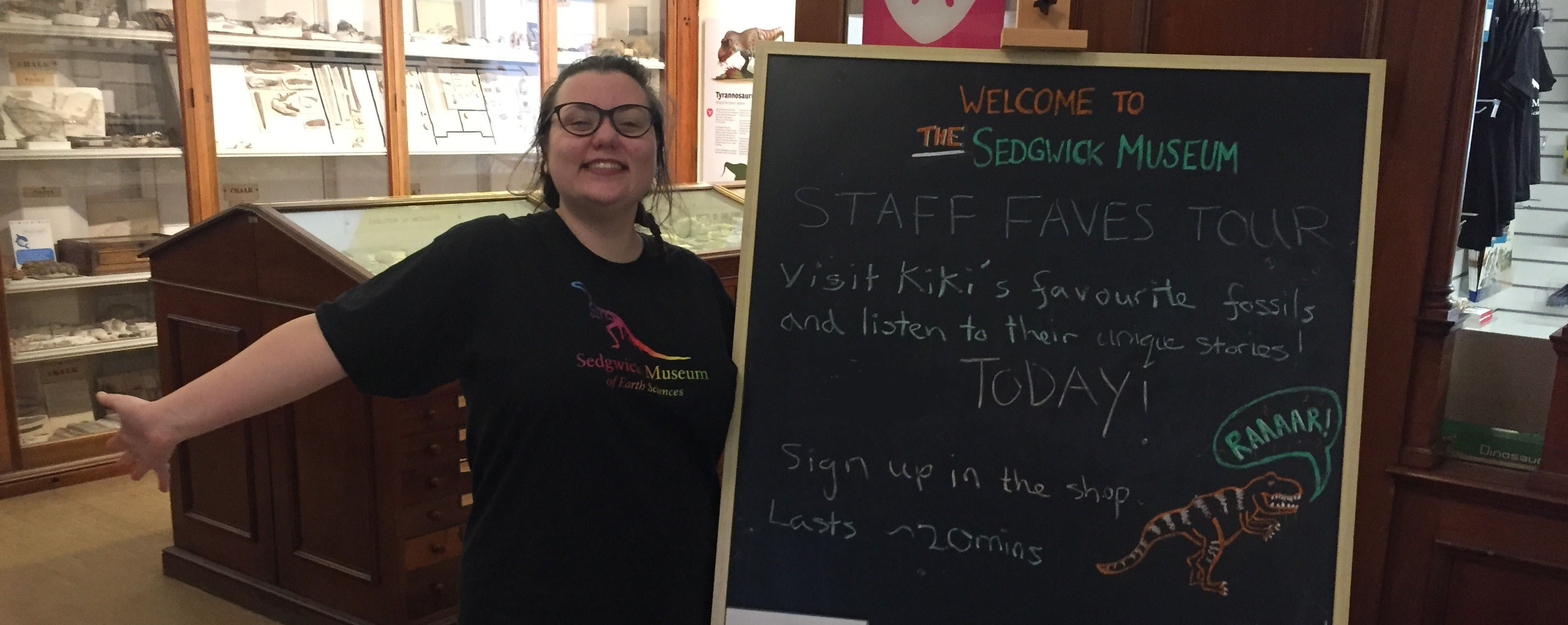 A member of museum staff smiles enthusiastically, arms outstretched, next to a large chalk board, announcing her Staff Favourites tour
