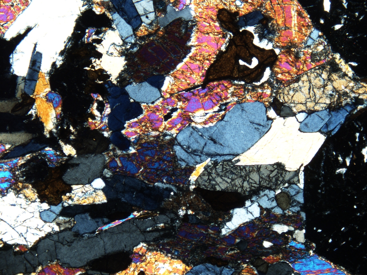 A 'thin section' of the mineral eclogite, as viewed down a petrographic microscope. It is a slice of rock, with areas of varied colours (different minerals), mainly black, grey, blue, pink, orange with small amounts of purple and yellow.