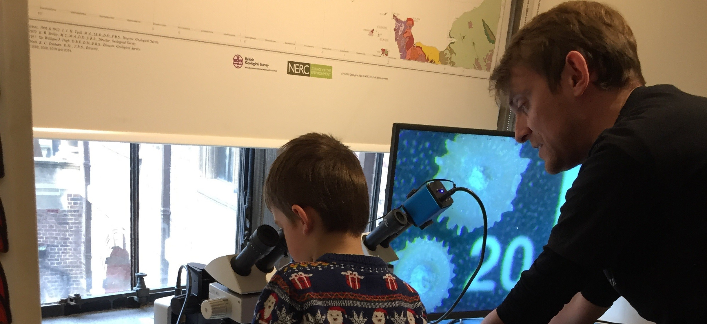 An adult helping a child to look down a microscope, with a star-shaped fossil on the screen attached to the microscope. This shows what the child is looking at.